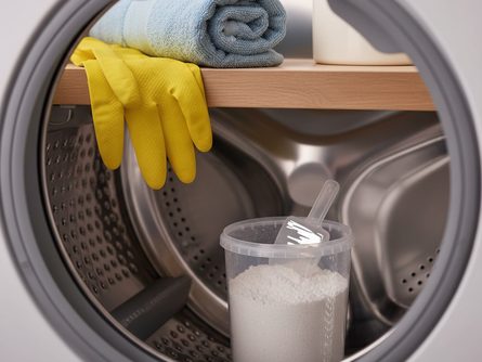 빈 세탁기 드럼 내부와 세탁조 클리너 분말이 담긴 투명한 용기, 고무 장갑과 마른 수건이 놓인 세탁실 선반